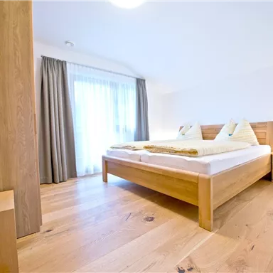 Ein modernes Schlafzimmer mit einem Holzbett und hellen Vorhängen. Der Fußboden ist aus hellem Holz, und es gibt genügend Tageslicht durch das Fenster.