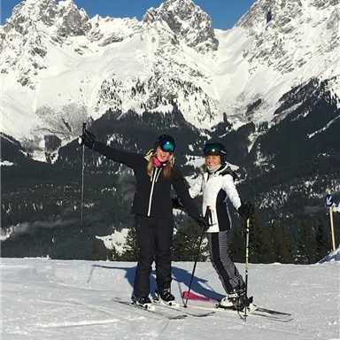 Zwei Skifahrerinnen stehen auf einem verschneiten Hang vor majestätischen Bergen. Der Himmel ist klar und blau, die Landschaft ist winterlich schön.