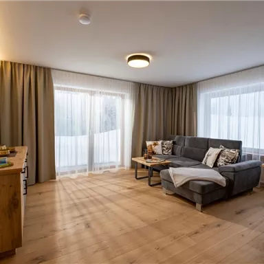 Ein modernes Wohnzimmer mit einem grauen Sofa und großen Fenstern. Der Raum hat Holzfußboden und helle Vorhänge, die viel Licht hereinlassen.