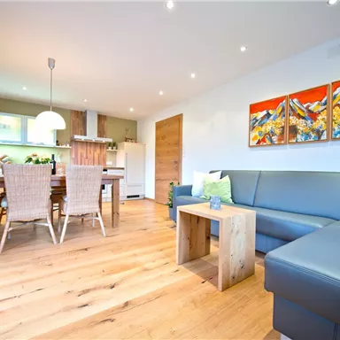 Ein helles Wohnzimmer mit einem modernen Sofa und einem Holztisch. An der Wand hängen bunte Gemälde, und der Raum ist freundlich und einladend.