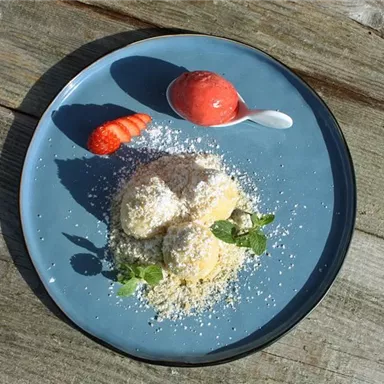 Eine elegante Dessert-Präsentation mit zwei Kugeln cremigem Eis, garniert mit Kokosraspeln, frischen Minzblättern und einer Erdbeere. Dazu ein Löffel mit fruchtigem Sorbet.