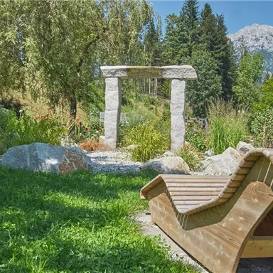 A beautiful garden with stone architecture and a deck chair. The landscape is green and surrounded by trees, with a view of the mountains.