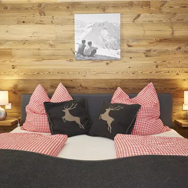 Ein gemütliches Schlafzimmer mit einem Holzwand und einem großen Bett. Die Kissen sind rot-weiß kariert und zeigen Hirschmotive, dazu gibt es warme Lampen.