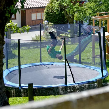 Ein Kind springt auf einem Trampolin im Garten. Im Hintergrund sind ein Spielplatz und Bäume zu sehen.