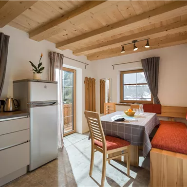 Eine helle und gemütliche Küche mit einem Esstisch und Holzdesign. Es gibt einen Kühlschrank und eine angenehme Sitzecke mit Fenstern, die Tageslicht hereinlassen.