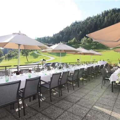 Eine einladende Terrasse mit lange gedeckten Tischen und Sonnenschirmen. Im Hintergrund erstreckt sich eine grüne Wiese und bewaldete Hügel.