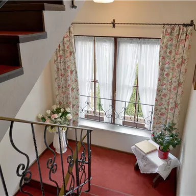 Ein einladender Flur mit einer Treppe und einem großen Fenster. Die Fenster sind mit weißen Vorhängen geschmückt, und eine Blumenarrangements sorgt für eine freundliche Atmosphäre.
