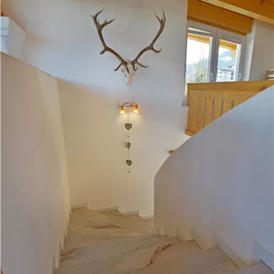 Eine helle Treppe mit einem Holzgeländer und einem Hirschgeweih an der Wand. Der Raum hat große Fenster, die natürliches Licht hereinlassen.