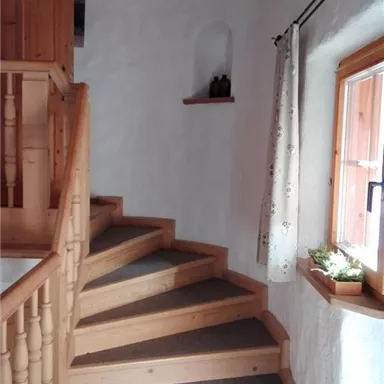 Eine helle Treppe aus Holz führt nach oben, umgeben von weißen Wänden. Ein Fenster lässt Tageslicht herein und bietet einen Blick nach draußen.