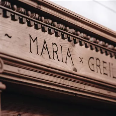 Ein kunstvoll geschnitzter Holzrahmen mit dem eingravierten Namen "Maria Greil". Die Struktur zeigt feine Details und verleiht dem Raum eine nostalgische Atmosphäre.