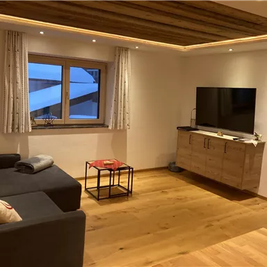 Ein gemütliches Wohnzimmer mit einem grauen Sofa und einem Fernseher an der Wand. Der Raum hat Holzdetails und große Fenster, die Tageslicht hereinlassen.