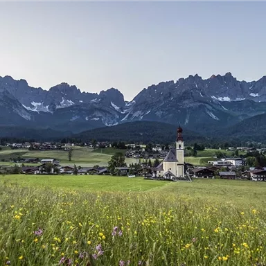 Ein malerisches Dorf mit einer Kirche im Vordergrund und beeindruckenden Bergen im Hintergrund. Die Wiese ist mit bunten Blumen bedeckt und strahlt eine ruhige Atmosphäre aus.
