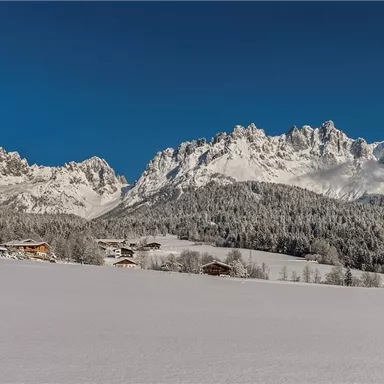 Eine malerische Winterlandschaft mit schneebedeckten Bergen und klar blauem Himmel. Im Vordergrund befinden sich traditionelle Hütten in einer ruhigen Umgebung.