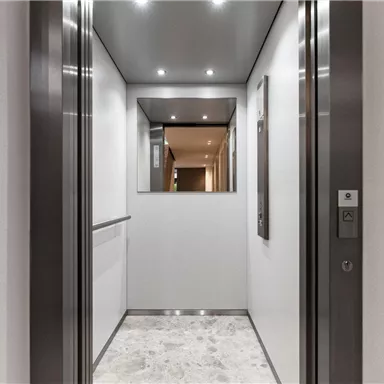A modern elevator with shiny walls and subtle lighting. The floor is made of light tiles and leads to an open door.