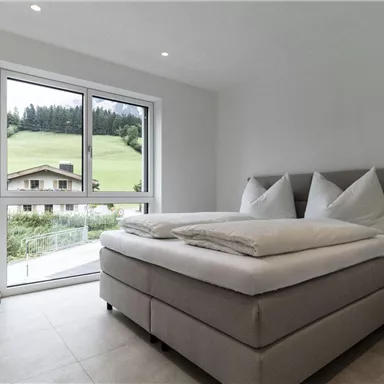 A modern bedroom with a large window that offers a view of green hills. The room is bright and welcoming with a comfortable bed and minimalist furnishings.