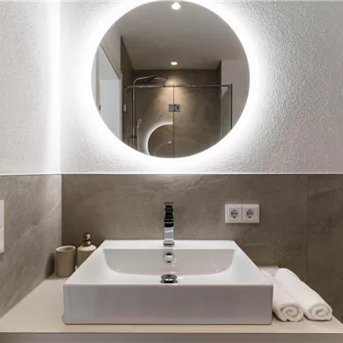 A modern bathroom with a stylish sink and a bright round mirror. The walls are in soft earthy tones.