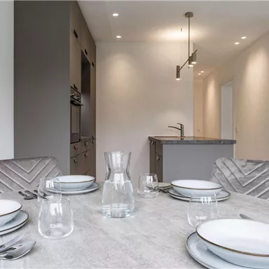 An elegant dining area with a round table set with dishes and glasses. In the background, a modern kitchen is visible.