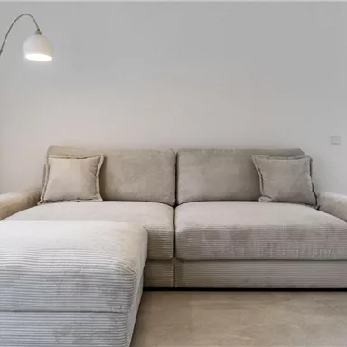 A modern, bright living room with a beige sofa and an elegant floor lamp. In the background, there are windows overlooking a green landscape and mountains.