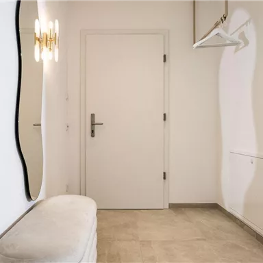 A bright hallway with a round mirror and a white bench. A coat hanger is hanging on the wall and the door is closed.