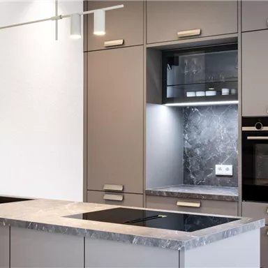 A modern kitchen with gray cabinets and an elegant countertop. It features a built-in oven and glass shelves with lighting.
