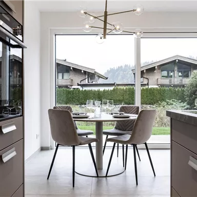A modern kitchen with a round table and upholstered chairs. The view overlooks a green property and mountainous architecture in the background.