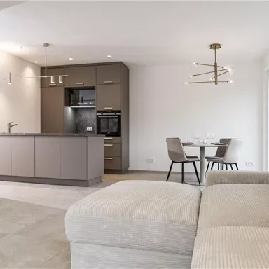 A modern kitchen with a dining area and a comfortable sofa. The room has bright walls and plenty of natural light.