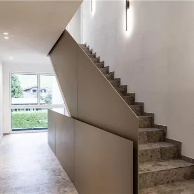 A modern entrance area with an elegant staircase. The walls are bright and designed with soft lighting accents.