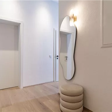 A bright hallway with modern white doors and an elegant mirror. The floor is made of light wood and there is a stylish seating area.