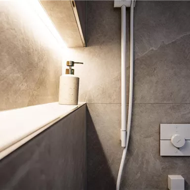 A modern bathroom with grey tiles and gentle lighting. An elegant soap dispenser stands on a shelf.