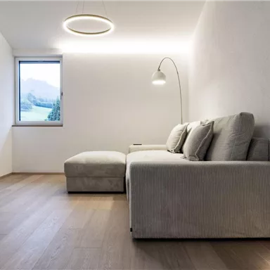 A modern living room with a beige sofa and a large window. A television is mounted on the wall, and there is a floor lamp for additional lighting.