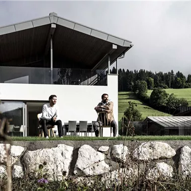 Two men are sitting relaxed on a terrace in front of a modern house. In the background, there is a green landscape with gentle hills and clouds.