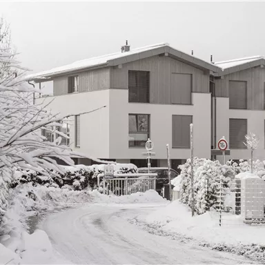 A modern house in the snow with a winding path. The surroundings are characterized by a wintry landscape that exudes tranquility.