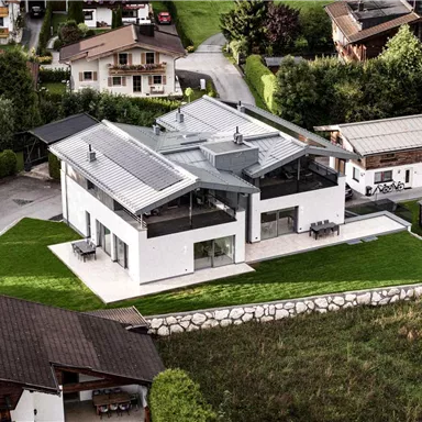 A modern house with a flat roof and large windows. The surroundings are green and well-maintained, with some other buildings nearby.