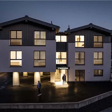 A modern building at night, illuminated with warm light. People are moving in front of the entrance and the surroundings are quiet.