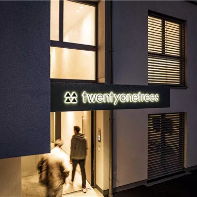 A modern building with the entrance of "twentyone trees". Two people are entering the building in evening lighting.