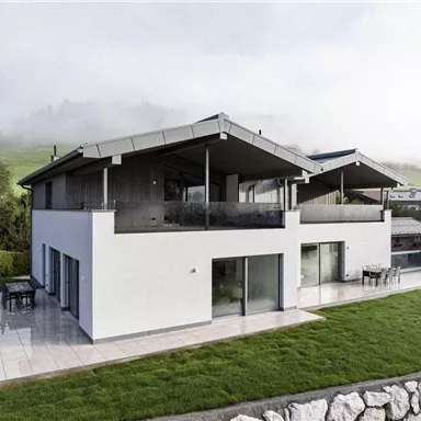 A modern house with large windows and a terrace area. The green lawn and the surrounding landscape create a peaceful atmosphere.