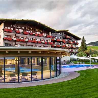 A modern hotel with a beautiful outdoor pool and well-kept garden. In the background, the mountains and a cloudy sky can be seen.