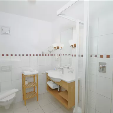 Ein modernes Badezimmer mit weißen Fliesen und einem klaren Duschbereich. Es gibt ein Holzregal mit Handtüchern und eine stilvolle Waschbecken-Einrichtung.