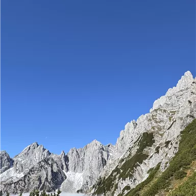 An impressive mountain landscape with steep rocks and green terrain. The sky is clear and blue, while clouds rest in the valley.
