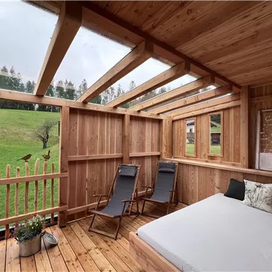 Ein gemütlicher Holzraum mit einem Bett und zwei Liegestühlen. Der Blick geht auf eine grüne Wiese und Bäume.