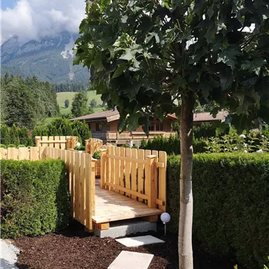 A stylish wooden bridge leads through a green hedge landscape. In the background, mountains and a blue sky can be seen.