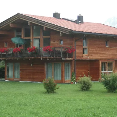 Ein gemütliches Holzhaus mit einem roten Dach und Balkonen, die mit Blumen geschmückt sind. Um das Haus herum befindet sich ein grüner Rasen und Berge im Hintergrund.