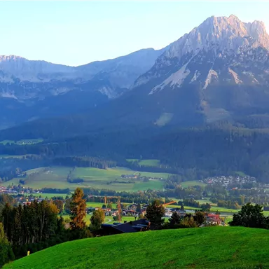 A breathtaking mountain landscape with green meadows and towering peaks. In the foreground, gentle hills and small houses are visible.