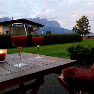 A table with two glasses of red drink and a candle stands in front of a picturesque mountainous landscape. In the background, trees and a building can be seen under a cloudy sky.