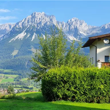 A picturesque mountain view with majestic peaks and green grass in the foreground. The image features a peaceful residential area with trees and a clear blue sky.