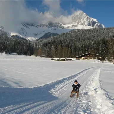 Ein Junge fährt mit einem Schlitten auf einem verschneiten Feld. Im Hintergrund sind Berge und ein Holzhaus zu sehen.