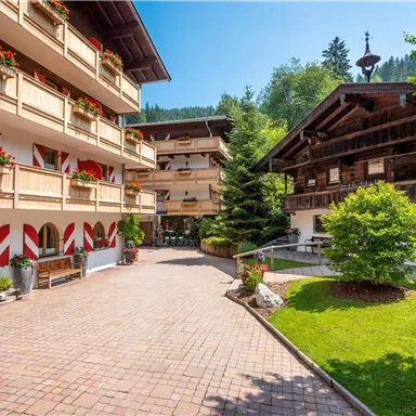 Ein malerischer Innenhof mit traditionellen Holzgebäuden und wunderschönen Blumen. Die grüne Landschaft und die umliegenden Bäume schaffen eine einladende Atmosphäre.