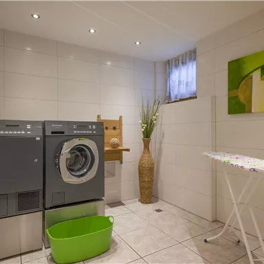 A modern laundry room with a washing machine and a dryer. An ironing station and a green laundry basket complete the equipment.