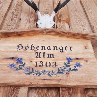 Ein Holzschild mit der Aufschrift "Höhenanger Ulm 1303" und einer Blumenornamentik. Darüber sind tierische Schädel mit Geweihen angebracht.