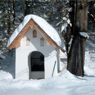 A small white house with a snow-covered roof stands in a snowy landscape. Surrounded by tall trees, the scene feels calm and idyllic.